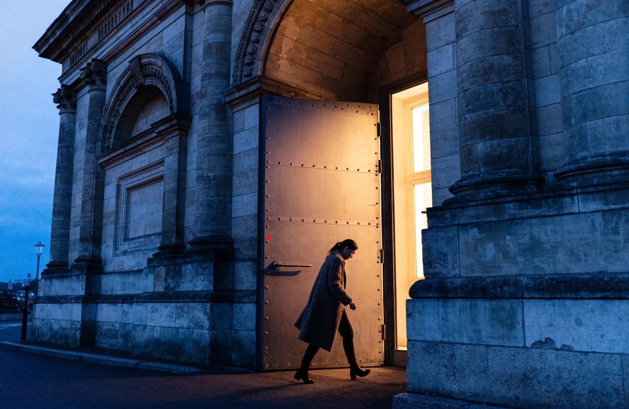 Treppenhaus eines Berliner Ministeriums in der Abenddämmerung, Marmorstufen und Messinghandlauf, eine Person im Mantel steigt hinauf, warmes Licht einer Wandlampe